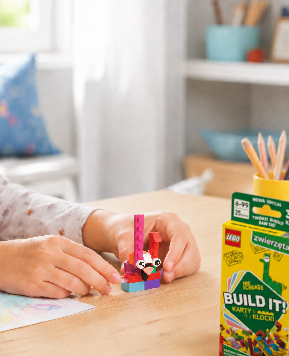 Dziecko buduje z klocków LEGO® kolorowego stworka obok pudełka BUILD IT!